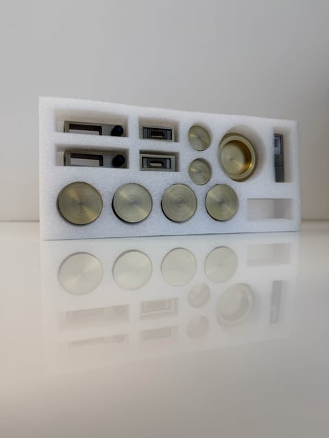 Organized foam storage tray containing various metallic speaker components including drivers, connectors, and metal discs arranged in compartments