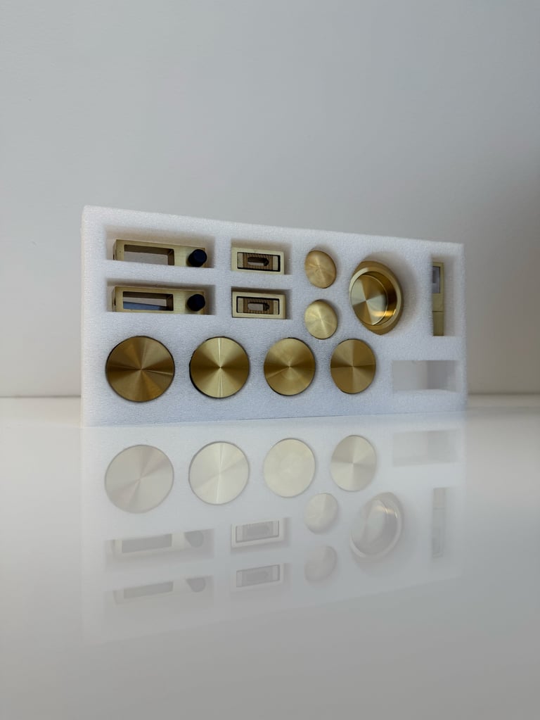 White foam display tray containing assorted brass and metal hardware components including cylindrical knobs, rectangular plates, circular knobs, and various metallic fixtures arranged in organized sections