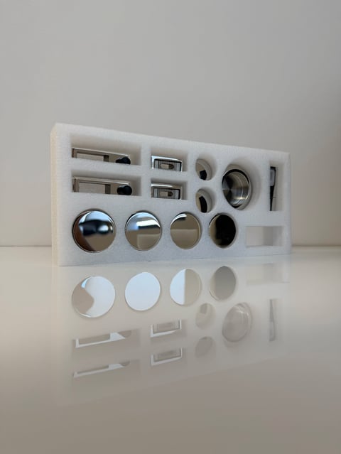 Organized collection of optical lenses, mirrors, and metal components arranged on white foam backing for product display or storage